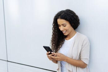 A woman leans against a wall, smiling while using her phone.