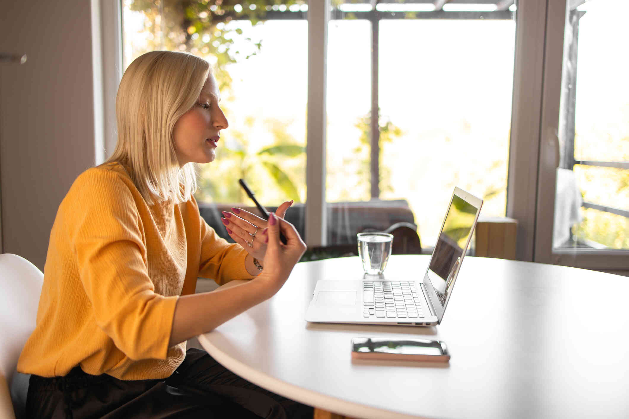 A woman in a yellow shirt sits at a table while talking to someone on her laptop