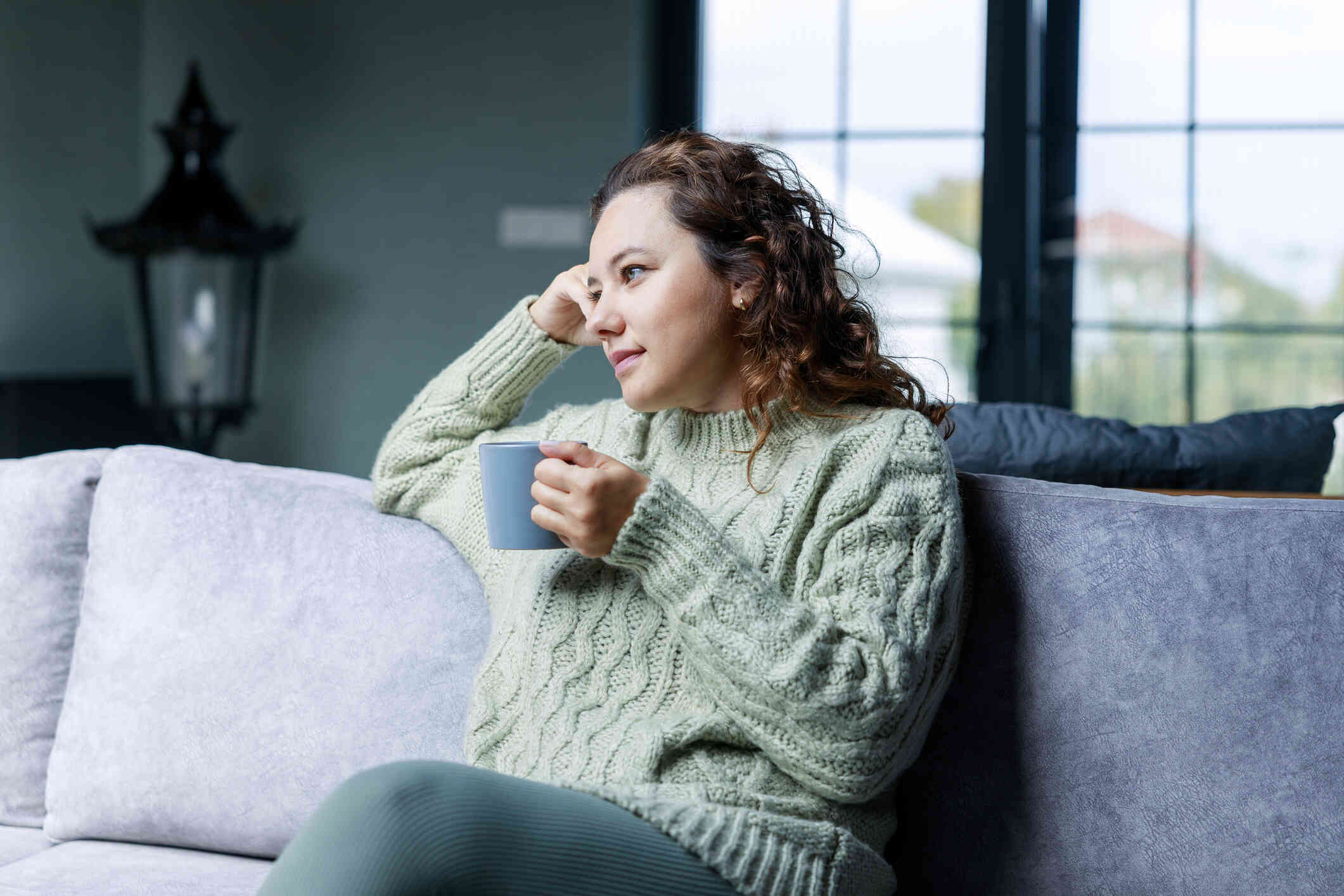 A woman has a neutral expression as she sits on a couch while wearing a large sweater and holds a coffee mug.
