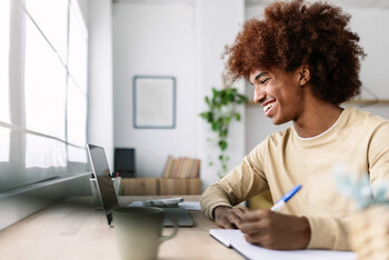 A man in a yellow shirt smiles at his laptop screen while taking notes.