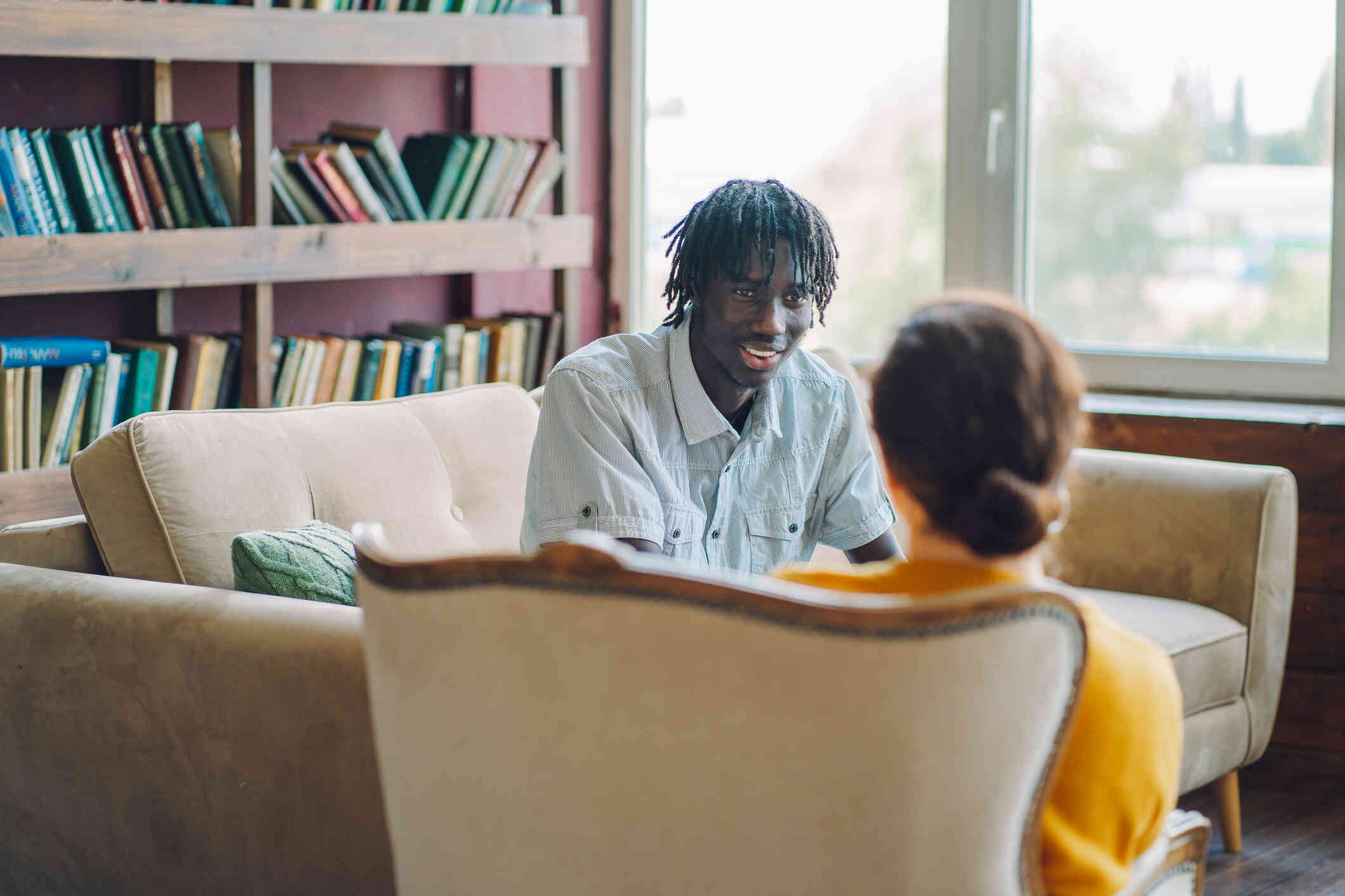 A man in a blue shirt sits on a couch while smiling and talking to a woman in an orange sweater