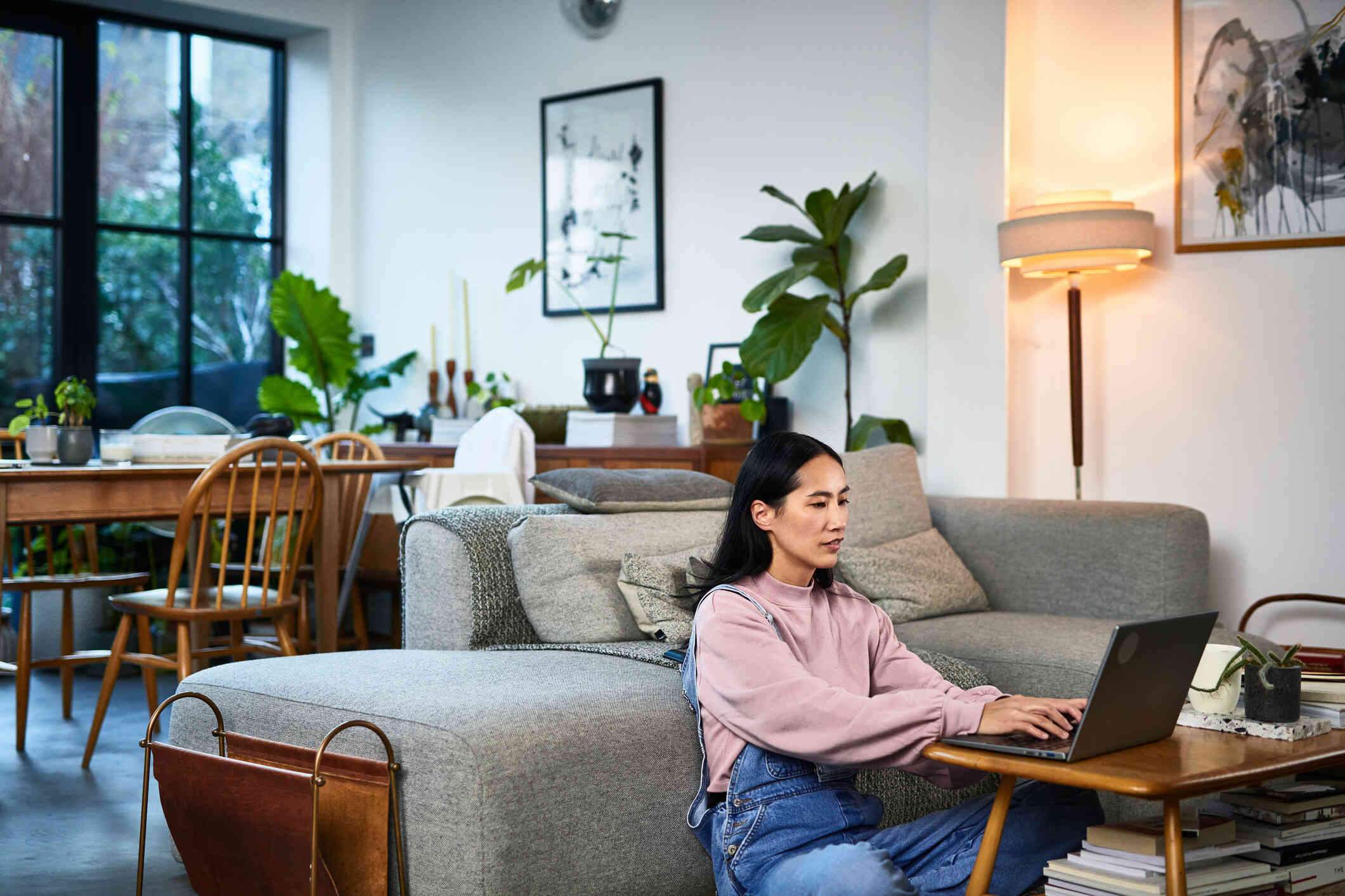 A woman wearing overalls sits on the floor in front of a couch in a house and looks at a laptop which is open on the coffee table in front of her.