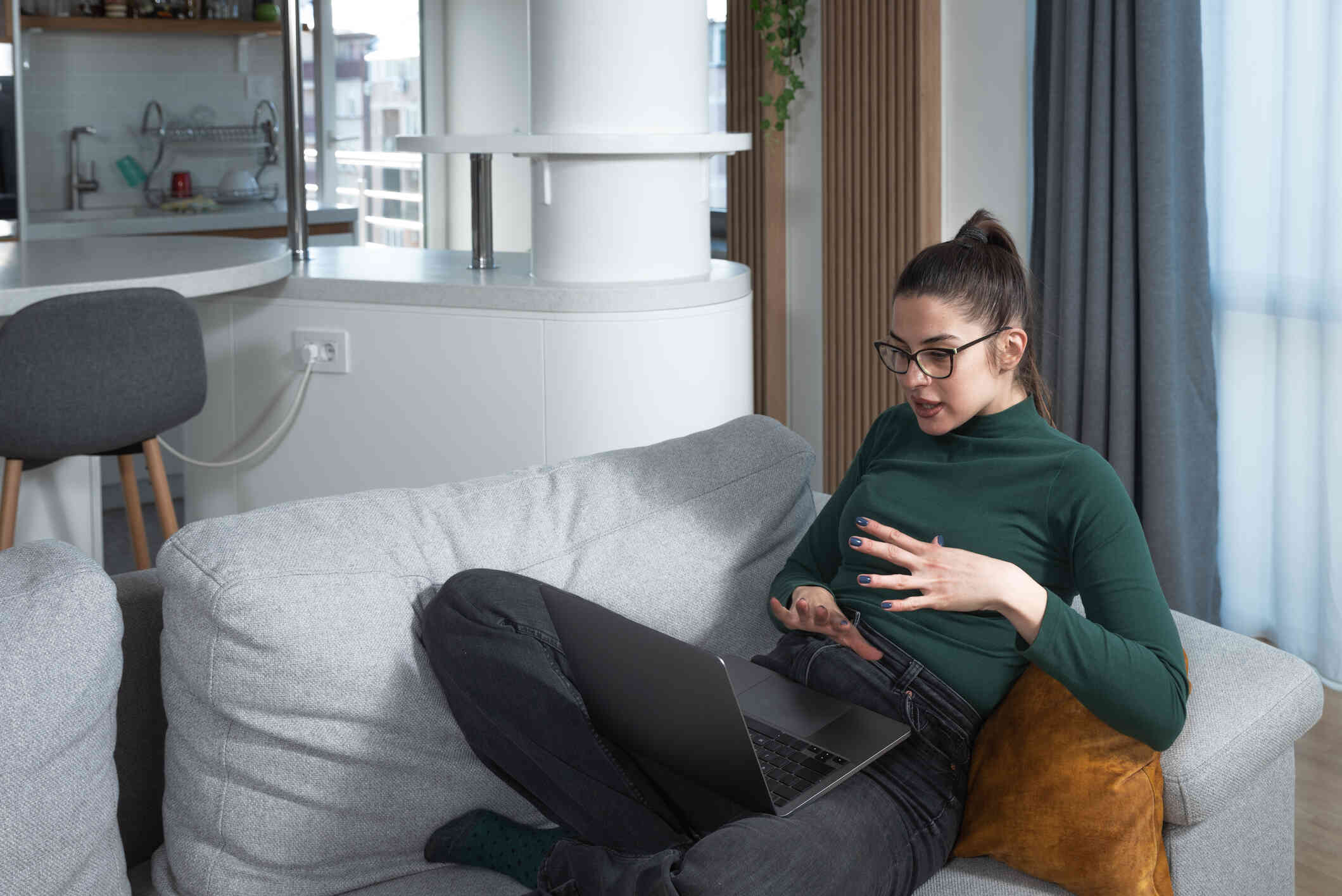 A woman in a green shirt sits on a couch while gesturing with her hands and talking to someone on her laptop