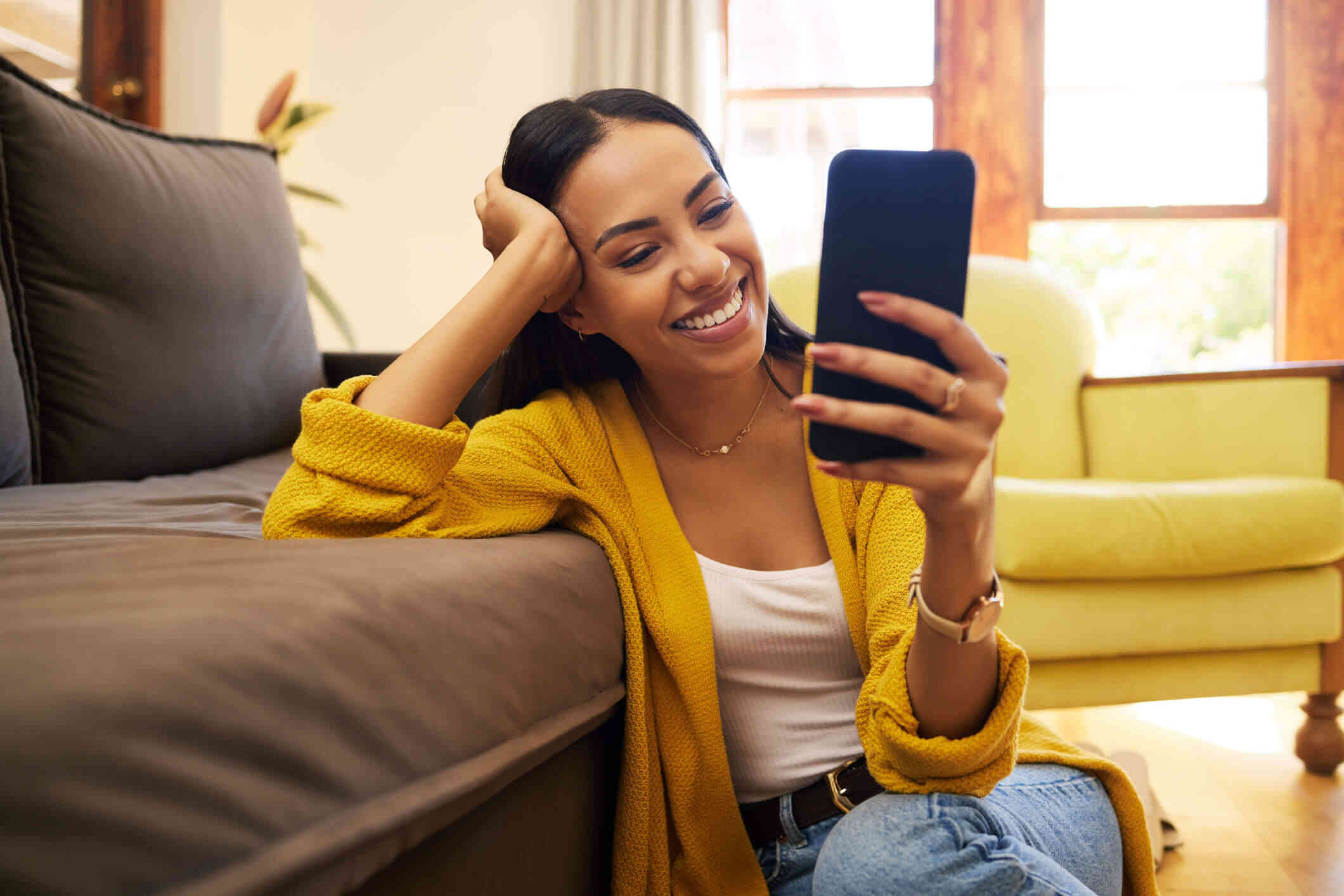 A women wearing a yellow sweater sits on the floor leaning on a couch while smiling at her phone