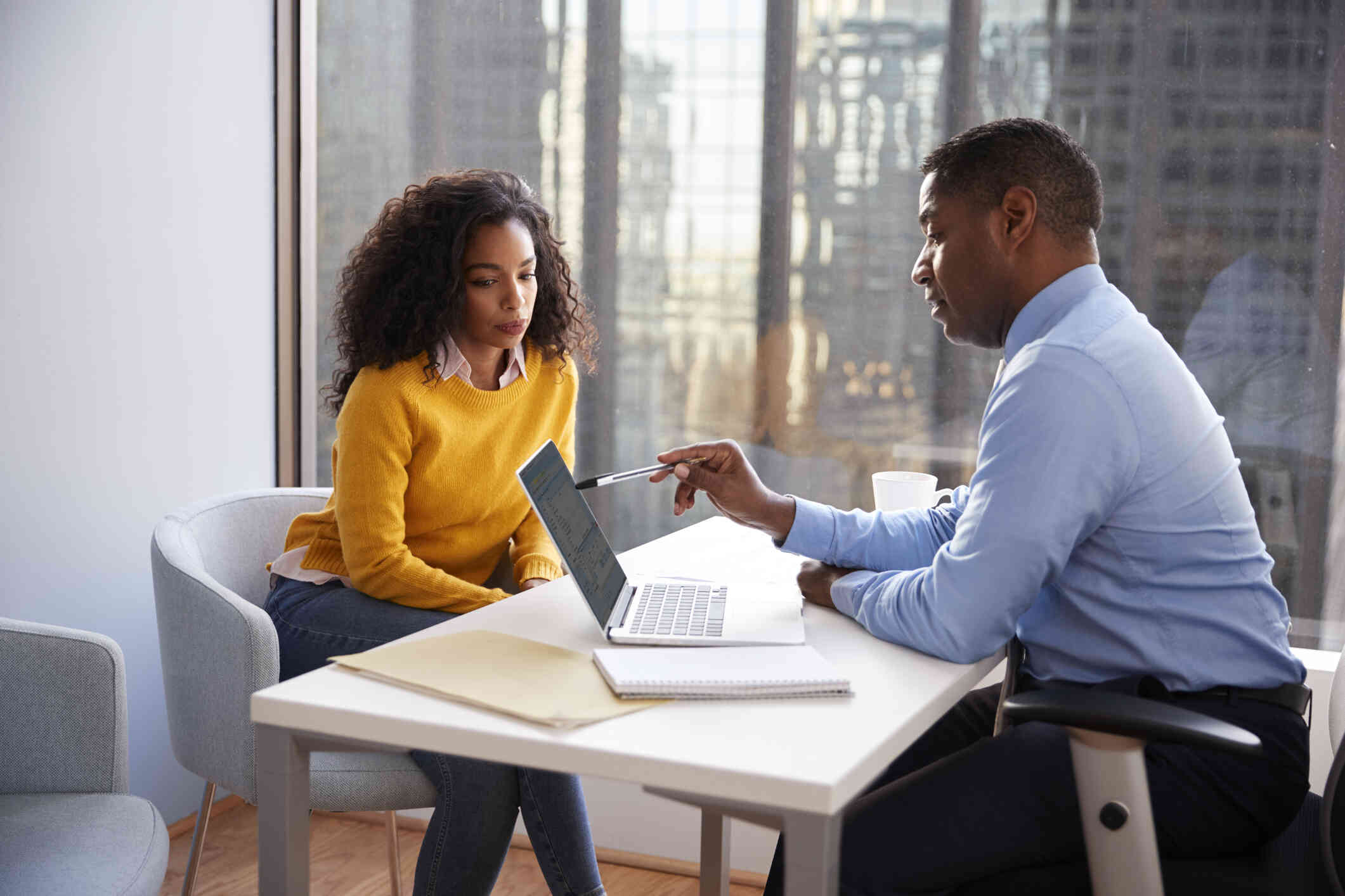 A man in a blue shirt sits with a woman in a yellow shirt as they discuss something on a laptop 