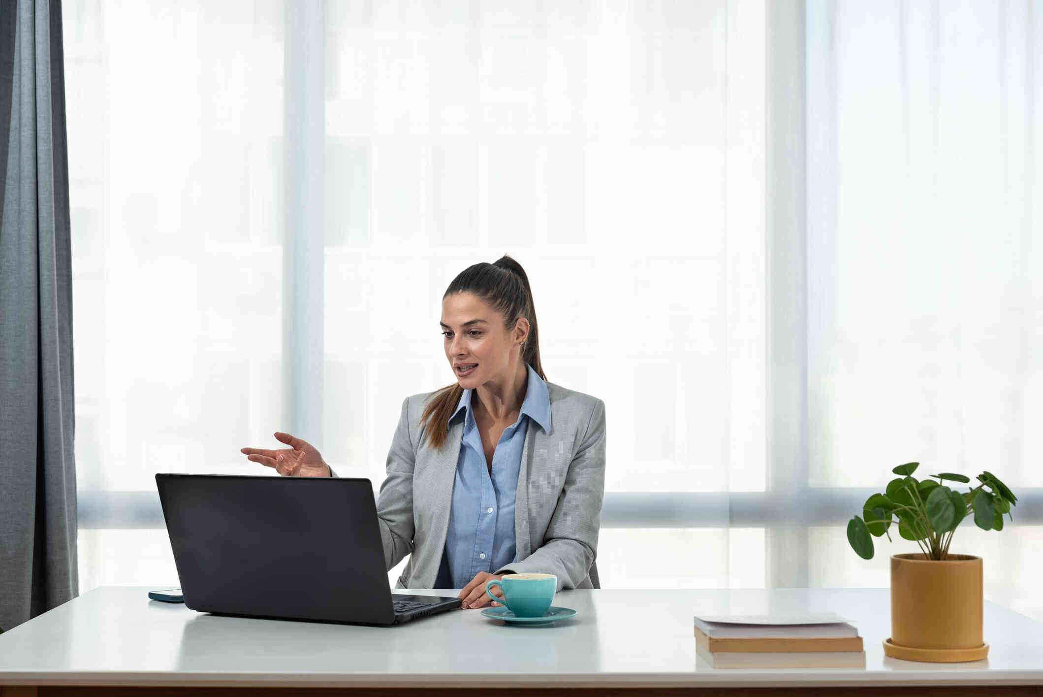 A woman in a blue shirt and green jacket sits at a desk while talking to someone on her laptop