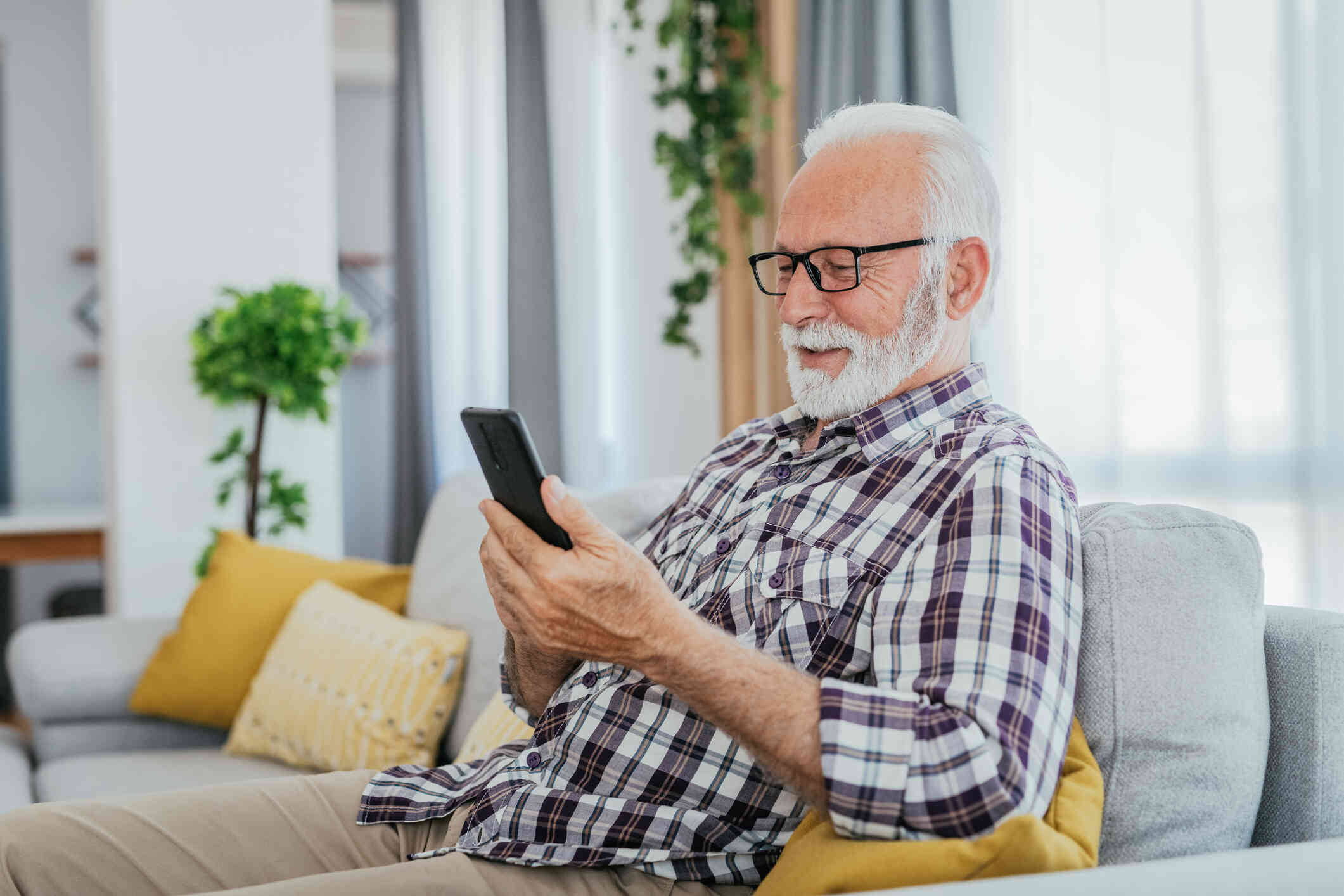 A man wearing glasses wearing a plaid shirt sits on a couch while smiling at his phone