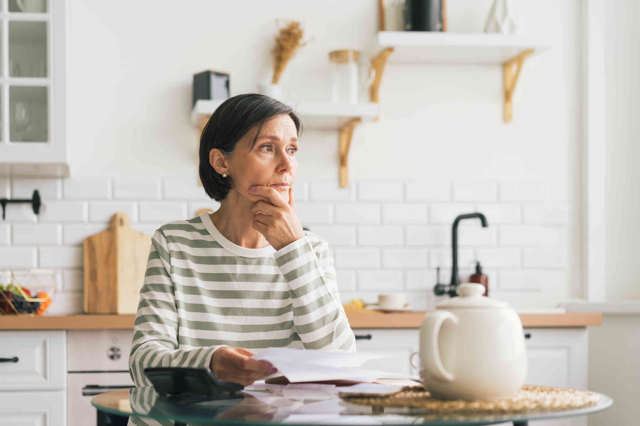 An older woman in striped sweater looks worried, seated at a kitchen table while holding documents.